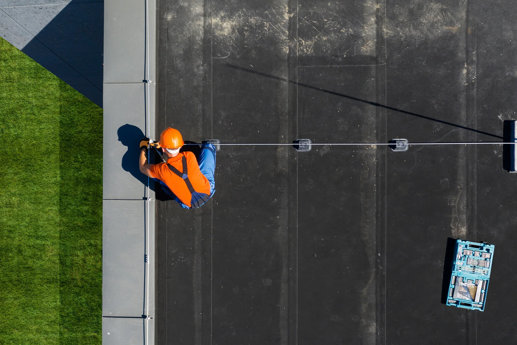 Overhead view of a roofing worker in high-visibility clothing installing a safety line along the edge of a flat commercial roof.