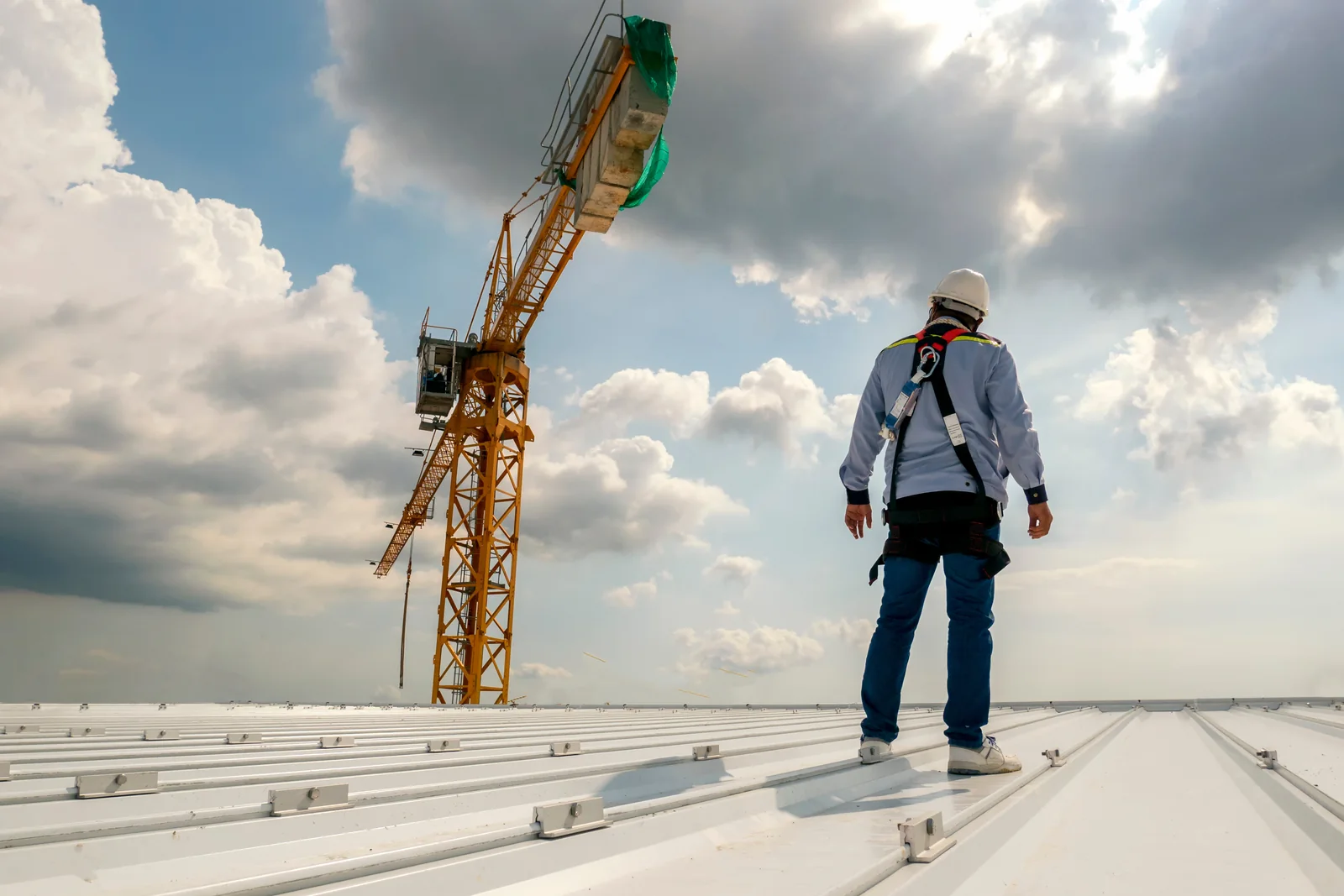 A roofing worker in a safety harness and hard hat standing on a large metal commercial roof, with a tower crane and dramatic sky behind.