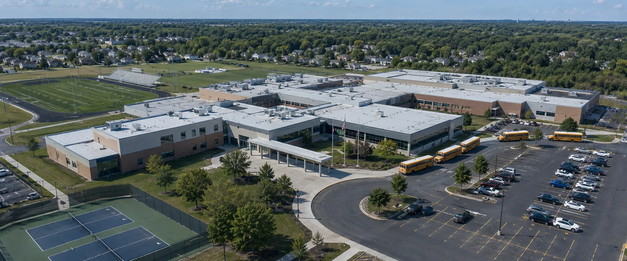 Aerial view of a large school campus with connected low-rise wings, a line of yellow school buses, a running track, tennis courts and a full car park.
